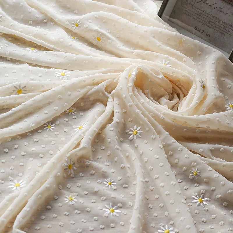 Gros plan sur le tissu mousseline plumetis brodé de marguerites au mètre de Longan Craft France, un tissu crème transparent orné de délicates marguerites blanches et jaunes, idéal pour les robes, les jupes, les chemisiers et la décoration.
