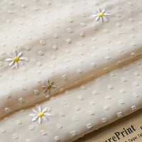 Un gros plan du tissu mousseline plumetis brodé de marguerites au mètre de Longan Craft France montre sa texture pointillée en relief et ses délicates marguerites blanches au centre jaune, le papier imprimé apparaissant en bas à droite.