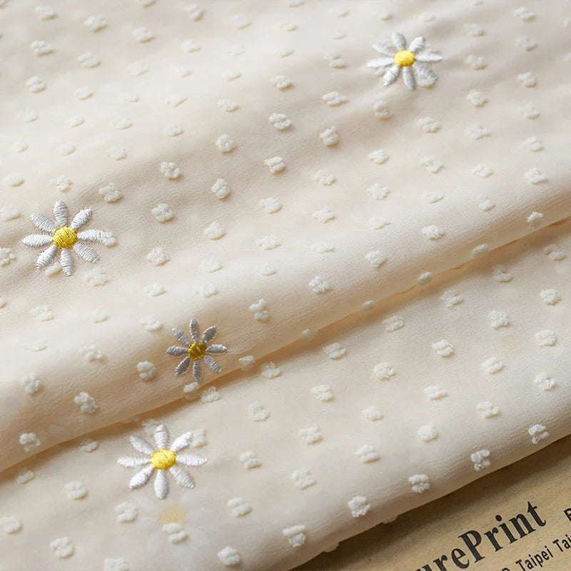 Un gros plan du tissu mousseline plumetis brodé de marguerites au mètre de Longan Craft France montre sa texture pointillée en relief et ses délicates marguerites blanches au centre jaune, le papier imprimé apparaissant en bas à droite.