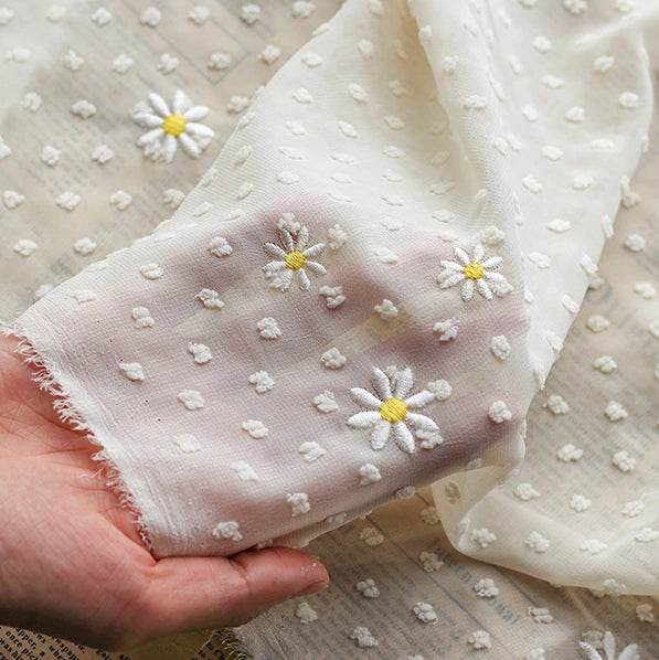 Une main tient le Tissu mousseline plumetis brodé de marguerites de Longan Craft France, un tissu transparent idéal pour les robes, les jupes, les chemisiers et la décoration. Ses broderies délicates et ses points en relief laissent deviner le contour des doigts.