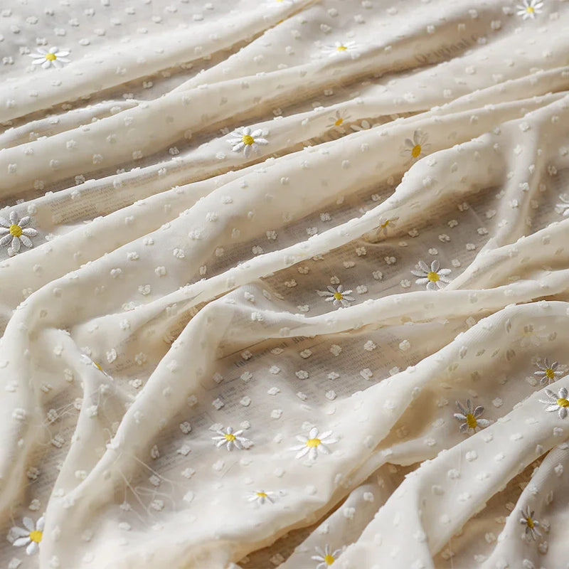 Un gros plan du tissu mousseline plumetis brodé de marguerites au mètre de Longan Craft France montre un tissu crème transparent à la texture délicate et légère, des points subtils et des marguerites blanches brodées, idéal pour les robes, les jupes, les chemisiers et la décoration.