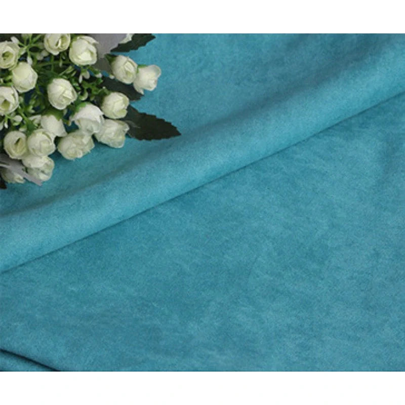 Un gros plan du Suédine unie embossée au mètre de Longan Craft France montre un daim synthétique doux et bleu clair à la texture subtile ; un petit bouquet de fleurs blanches et de feuilles vertes apparaît dans le coin supérieur gauche.