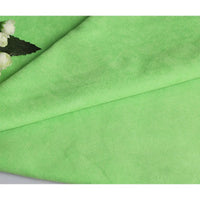 Un gros plan de Suédine unie embossée au mètre par Longan Craft France montre sa texture de daim synthétique lisse, vert clair, plié, avec des fleurs blanches et des feuilles vertes dans le coin supérieur gauche. Un tissu haut de gamme idéal pour la confection de vêtements.