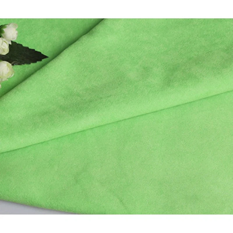 Un gros plan de Suédine unie embossée au mètre par Longan Craft France montre sa texture de daim synthétique lisse, vert clair, plié, avec des fleurs blanches et des feuilles vertes dans le coin supérieur gauche. Un tissu haut de gamme idéal pour la confection de vêtements.