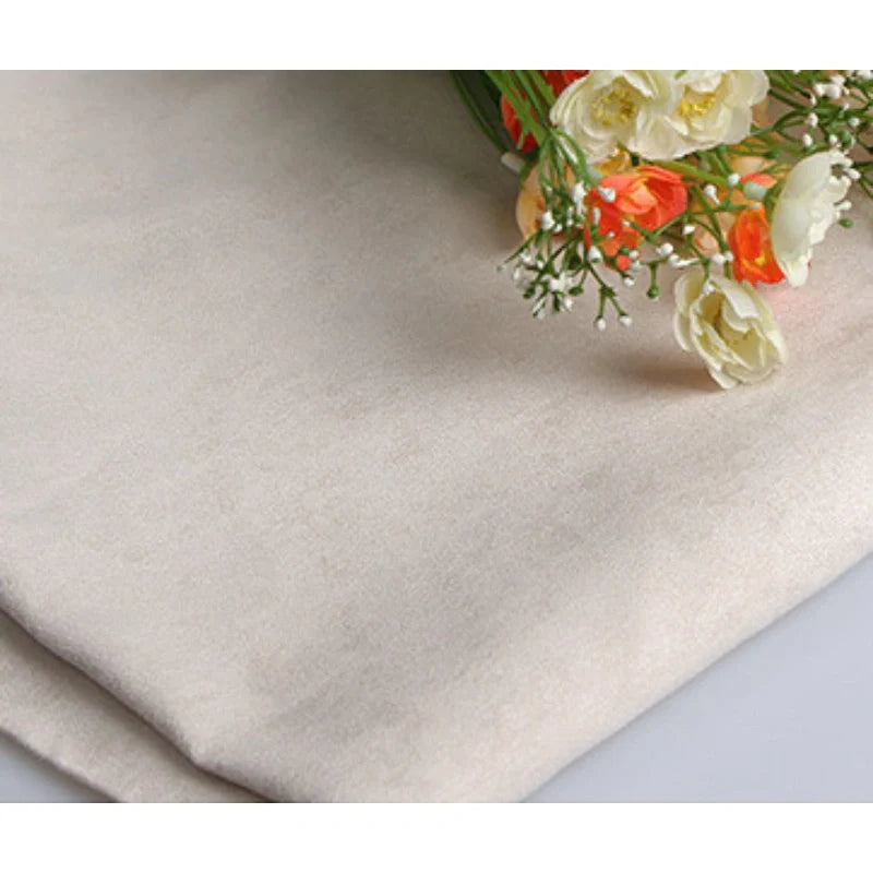 Gros plan sur le Suédine unie embossée au mètre de Longan Craft France en beige clair, plié avec des fleurs artificielles crème et orange sur le dessus. Ce daim synthétique de haute qualité est idéal pour la confection de vêtements et de coussins.