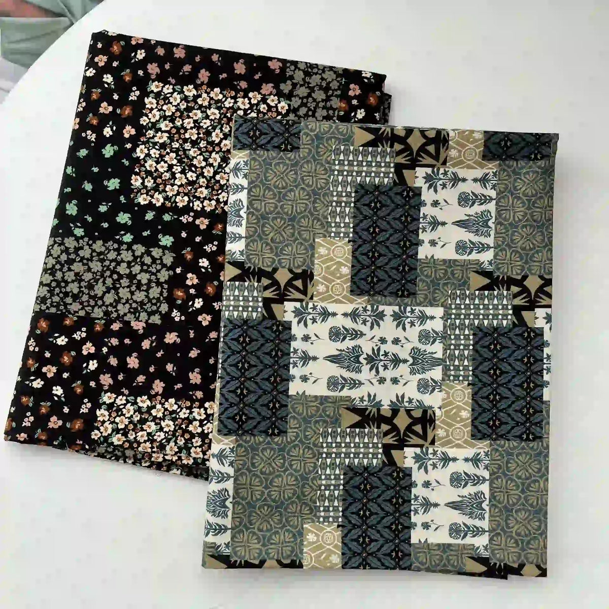 Deux tissus pliés sur une surface blanche : l'un est un Linon de coton imprimé floral ethnique de Longan Craft France sur fond noir, l'autre est un patchwork géométrique dans des tons verts, bleus, beiges et noirs.