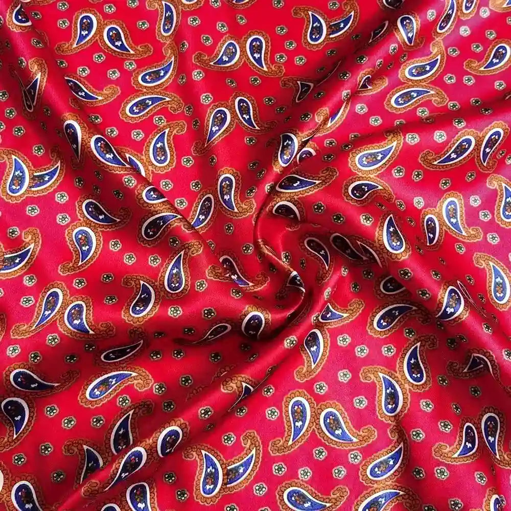 Un gros plan du Tissu satin polyester imprimé paisley au mètre de Longan Craft France montre son motif cachemire coloré en bleu, jaune et blanc sur du satin rouge, avec des plis doux et des ombres qui donnent au tissu un aspect élégant.