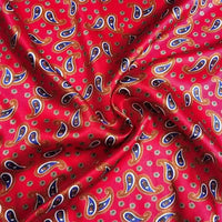 Un gros plan du Tissu satin polyester imprimé paisley au mètre de Longan Craft France montre son motif cachemire coloré en bleu, jaune et blanc sur du satin rouge, avec des plis doux et des ombres qui donnent au tissu un aspect élégant.