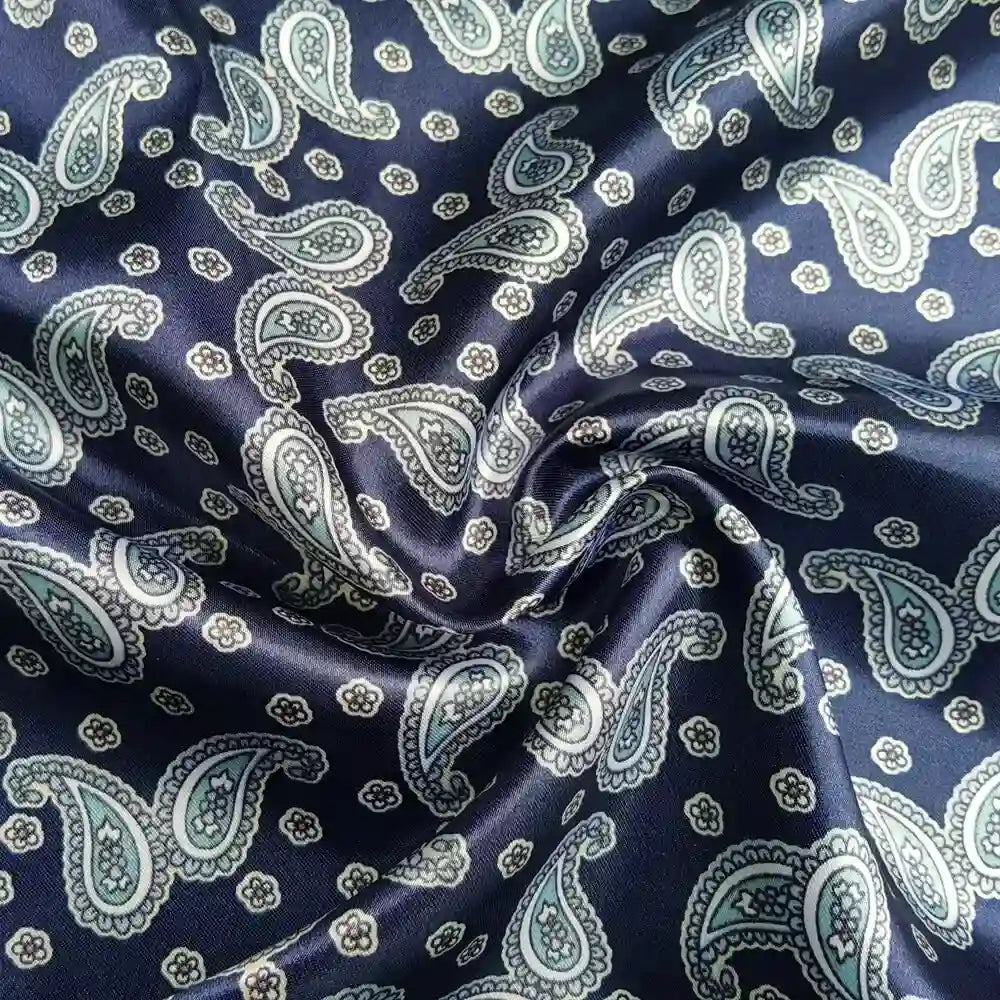 Un gros plan du tissu satin polyester imprimé paisley au mètre de Longan Craft France montre des motifs cachemire bleu, blanc et bleu clair avec des plis visibles dans l'élégant tissu satin.