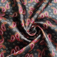 Gros plan sur le Tissu satin polyester imprimé paisley de Longan Craft France, noir brillant avec motifs cachemire rouges et bruns et petites fleurs blanches, drapé en plis doux.