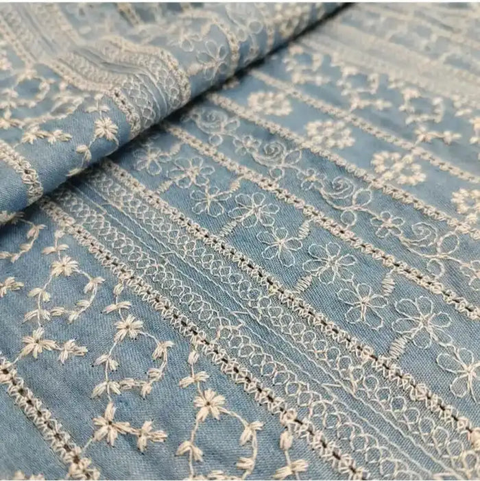 Un gros plan de "Tissu en coton brodé floral au mètre" de Longan Craft France montre un tissu denim léger avec des broderies florales et géométriques blanches complexes, astucieusement présentées en plis superposés, idéales pour les robes et les chemisiers.