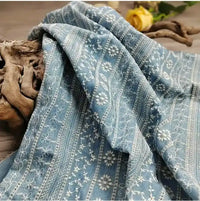 Le Tissu en coton brodé floral au mètre de Longan Craft France - un tissu brodé léger de style denim - apparaît drapé sur du bois flotté, avec une rose jaune et des touches de bois en arrière-plan.