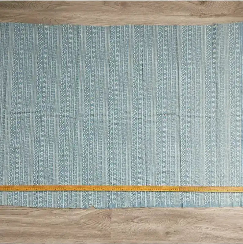 Un morceau de Tissu en coton brodé floral au mètre de Longan Craft France est posé à plat sur un plancher en bois, un mètre ruban jaune étant placé horizontalement le long du bord inférieur du tissu.