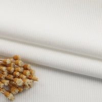 Un morceau plié de tissu velours côtelé à fines côtes au mètre de Longan Craft France, en blanc, est présenté avec un bouquet de fleurs séchées beiges sur le dessus. La texture côtelée et le fond assorti mettent en valeur ce tissu pour les canapés ou les vêtements.