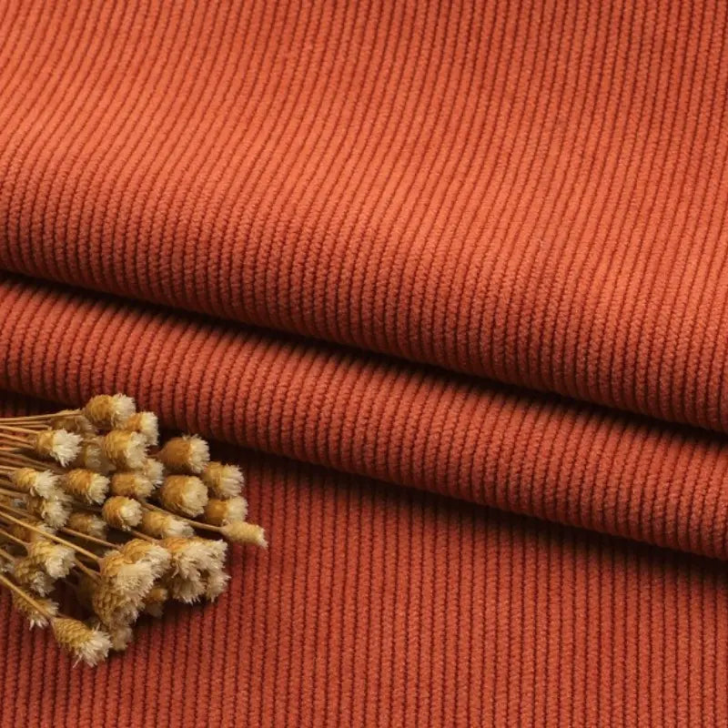 Gros plan sur le Tissu velours côtelé à fines côtes au mètre de Longan Craft France, de couleur rouille, soigneusement plié et surmonté d'un petit paquet de fleurs beiges séchées - idéal pour les canapés, les vêtements ou les pantalons.