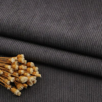 Gros plan sur un pli de Tissu velours côtelé gris foncé à fines côtes au mètre de Longan Craft France, avec des fleurs jaunes séchées sur le dessus. La texture nervurée et le motif linéaire de ce tissu pour canapé, vêtements, pantalons sont clairement visibles.