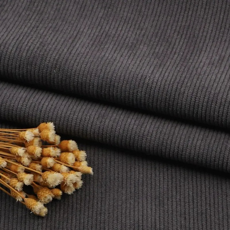 Gros plan sur un pli de Tissu velours côtelé gris foncé à fines côtes au mètre de Longan Craft France, avec des fleurs jaunes séchées sur le dessus. La texture nervurée et le motif linéaire de ce tissu pour canapé, vêtements, pantalons sont clairement visibles.