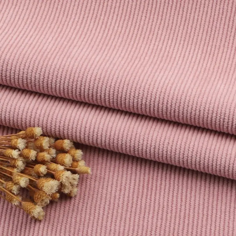 Le tissu velours côtelé à fines côtes au mètre de Longan Craft France est empilé sur une surface, avec des fleurs séchées dans un coin. La texture de ce velours uni, idéal pour les canapés et les vêtements, et les détails des fleurs sont clairement visibles.