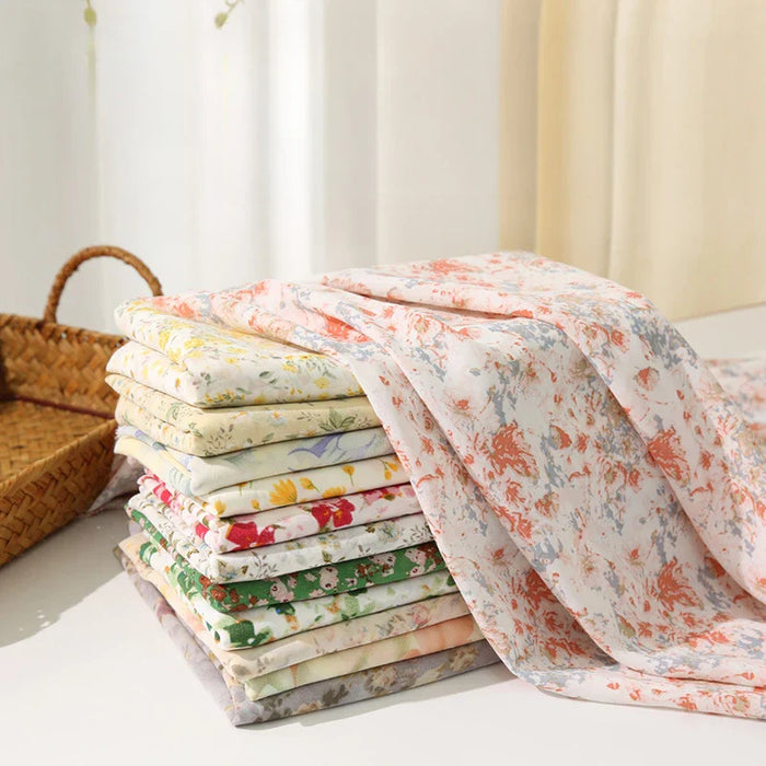 Une pile de Tissu mousseline à fleurs au mètre - mousseline de polyester imprimée pour l'habillement - aux motifs floraux pastel de Longan Craft France est soigneusement pliée sur une surface blanche à côté d'un panier, avec un rideau moelleux en arrière-plan.