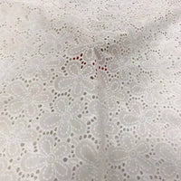 Un gros plan du Tissu œillet brodé floral au mètre de Longan Craft France montre un tissu blanc 100 % coton avec un délicat motif floral à œillets, de petites découpes rondes et de subtils tons rouges visibles sous sa texture légère et aérée.