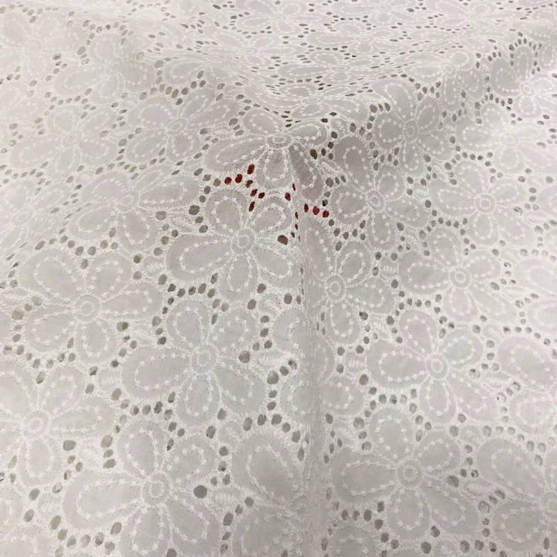 Un gros plan du Tissu œillet brodé floral au mètre de Longan Craft France montre un tissu blanc 100 % coton avec un délicat motif floral à œillets, de petites découpes rondes et de subtils tons rouges visibles sous sa texture légère et aérée.