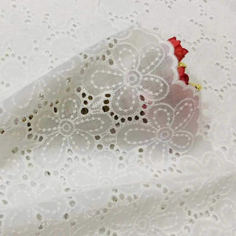 Le Tissu œillet brodé floral au mètre de Longan Craft France, léger et aérien à motifs fleuris, est délicatement posé sur des fleurs rouges et jaunes artificielles, visibles sous la matière transparente.