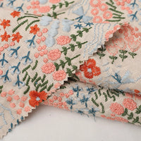 Gros plan sur le Tissu brodé floral au mètre | Tissu coton lin brodé fleurs et plantes de Longan Craft France, qui présente des broderies complexes orange, bleues et vertes et des bords rosés - idéal pour la couture ou la décoration intérieure.