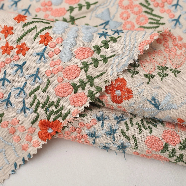 Gros plan sur le Tissu brodé floral au mètre | Tissu coton lin brodé fleurs et plantes de Longan Craft France, qui présente des broderies complexes orange, bleues et vertes et des bords rosés - idéal pour la couture ou la décoration intérieure.