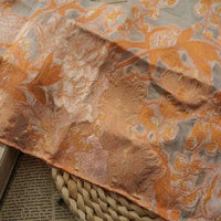 Le Tissu organza brodé floral au mètre de Longan Craft France est un délicat tissu transparent avec une broderie florale orange, présentée sur une surface tissée et recouvrant partiellement un journal - idéal pour les robes et les usages décoratifs.