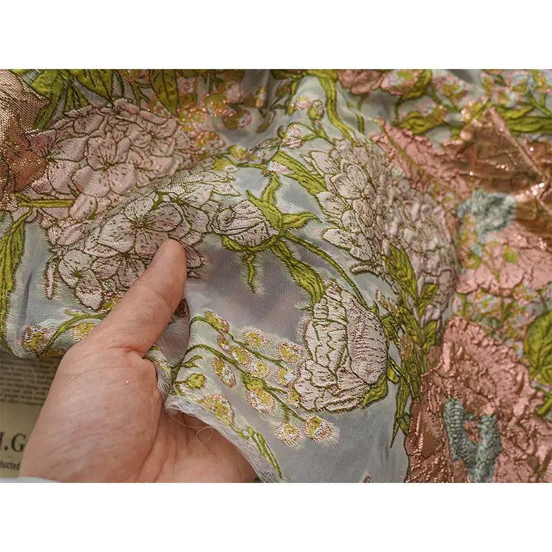 Une main tient le Tissu organza brodé floral au mètre de Longan Craft France, un organza semi-transparent pour les robes avec des broderies vertes, roses et dorées ; à travers le tissu délicat, un journal est partiellement visible.