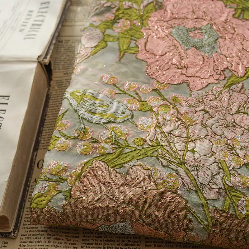 Un livre recouvert de tissu et orné d'un motif floral et d'oiseaux brodés, inspiré du Tissu organza brodé floral au mètre de Longan Craft France, est posé sur un journal ouvert à côté d'un autre livre aux pages légèrement gondolées.