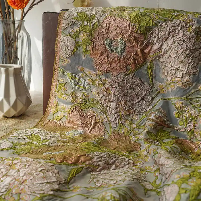 Un morceau de Tissu organza brodé floral au mètre de Longan Craft France est drapé sur une table. L'organza transparent présente une broderie florale complexe rose, verte et dorée, avec un vase géométrique blanc et des fleurs orange en arrière-plan.