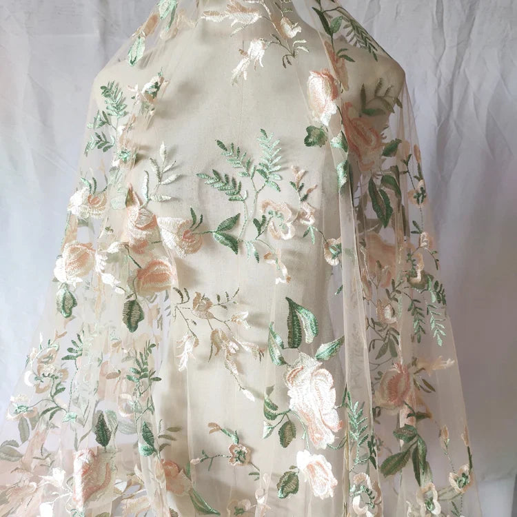 Le Tulle brodé floral au mètre de Longan Craft France est un tissu brodé de fleurs rose pâle et de feuilles vertes sur un fond clair, idéal pour la couture des robes de mariée.