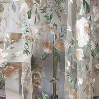 Des rideaux en Tulle brodé floral au mètre de Longan Craft France, aux motifs brodés rose pâle, vert et crème, sont suspendus devant un lampadaire. Une table avec un bol beige et une petite plante est partiellement visible derrière.