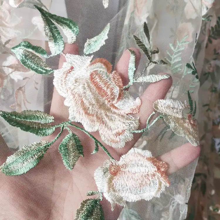 Une main présente le Tulle brodé floral au mètre de Longan Craft France, un tissu transparent brodé de fleurs pastel et de feuilles vertes, parfait pour la couture de robes de mariée. La matière translucide laisse apparaître la lumière et la main.