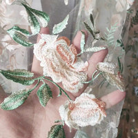 Une main présente le Tulle brodé floral au mètre de Longan Craft France, un tissu transparent brodé de fleurs pastel et de feuilles vertes, parfait pour la couture de robes de mariée. La matière translucide laisse apparaître la lumière et la main.