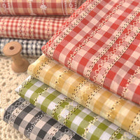 Des piles de "Tissu popeline fleurie et vichy au mètre" de Longan Craft France en rouge, rose, jaune, vert et noir avec des bordures en dentelle - fabriquées en coton polyester - sont disposées avec art à côté d'une bobine en bois sur une surface crochetée.