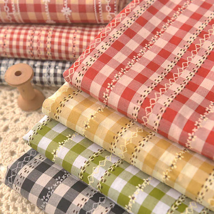 Des piles de "Tissu popeline fleurie et vichy au mètre" de Longan Craft France en rouge, rose, jaune, vert et noir avec des bordures en dentelle - fabriquées en coton polyester - sont disposées avec art à côté d'une bobine en bois sur une surface crochetée.