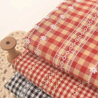 Trois pièces pliées de Tissu popeline fleurie et vichy de Longan Craft France à carreaux rouges, jaunes et noirs avec broderie blanche sont empilées sur de la dentelle, avec une bobine en bois à côté d'elles. Tissu élégant en coton-polyester pour l'habillement féminin.