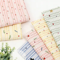 Plusieurs pièces de Tissu pour patchwork vichy au mètre et à motif botanique Longan Craft France, en polyester confortable, sont disposées sur une surface blanche dans des tons pastel, avec une petite plante verte visible en bas à gauche.