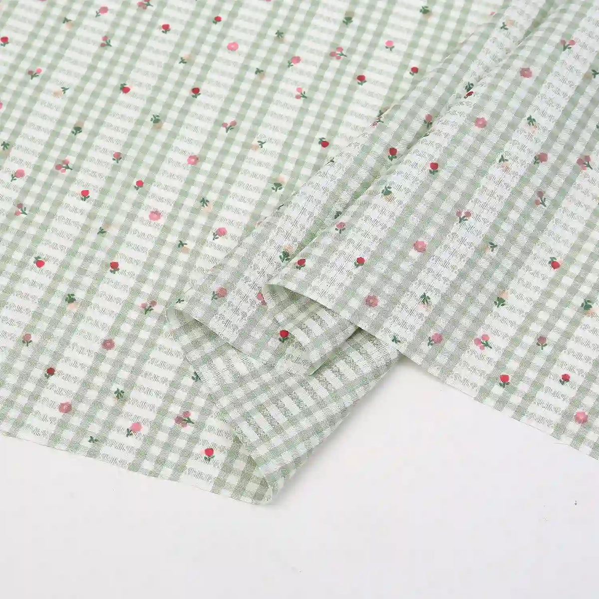 Un tissu Longan Craft France patchwork vichy au mètre en vichy vert clair et blanc avec des motifs botaniques roses et rouges, soigneusement plié, repose sur une surface blanche. Tissu en polyester, idéal pour les tenues décontractées et les projets créatifs.