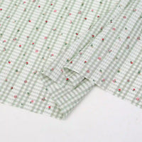 Un tissu Longan Craft France patchwork vichy au mètre en vichy vert clair et blanc avec des motifs botaniques roses et rouges, soigneusement plié, repose sur une surface blanche. Tissu en polyester, idéal pour les tenues décontractées et les projets créatifs.