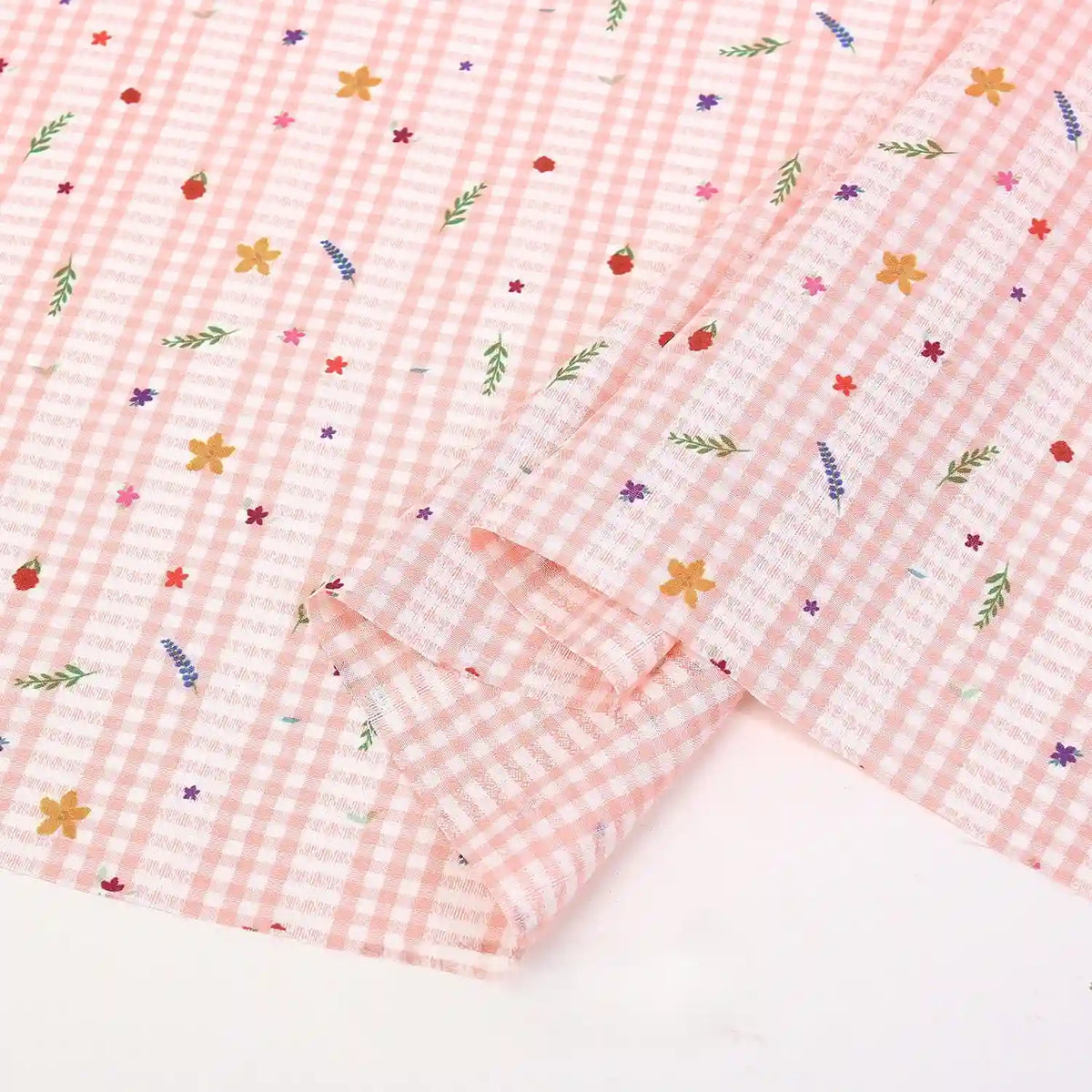 Un morceau du tissu pour patchwork vichy au mètre de Longan Craft France, en polyester confortable, arbore un motif botanique rose clair et blanc avec des fleurs et feuilles colorées, partiellement plié pour révéler sa texture.