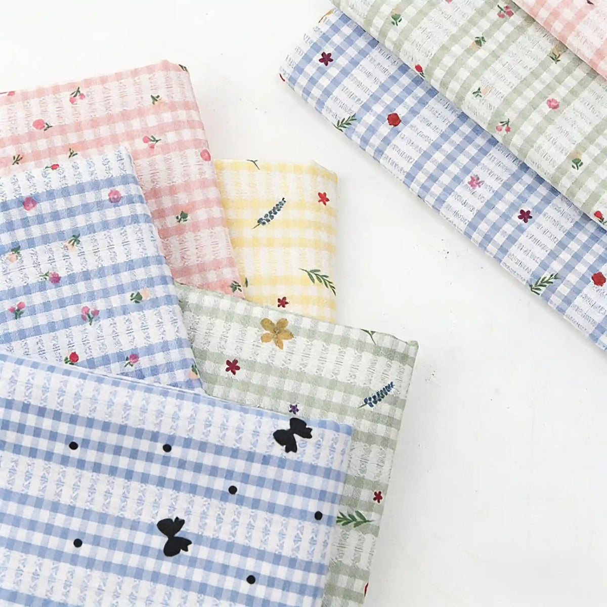 Plusieurs pièces du tissu patchwork vichy au mètre en polyester de Longan Craft France sont disposées sur une surface blanche, mettant en valeur de confortables motifs à carreaux pastel avec des motifs botaniques et floraux, idéaux pour les vêtements décontractés.