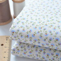 Deux pièces de Tissu linon de coton fleuri de Longan Craft France, présentant un imprimé floral bleu et rouge, sont présentées à côté d'une règle en bois et de deux bobines de fil (l'une marron, l'autre blanche). Idéal pour les vêtements et les doublures.