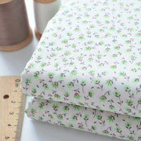 Deux morceaux pliés de Tissu linon de coton fleuri de Longan Craft France, présentant un petit motif floral vert, sont empilés à côté d'une règle en bois et de deux bobines de fil (marron et blanc) sur une surface blanche.
