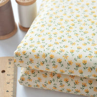 Deux morceaux pliés de Tissu linon de coton fleuri au mètre de Longan Craft France, avec un voile de coton floral jaune, sont placés à côté d'une règle en bois et de deux bobines de fil, l'une marron et l'autre crème.