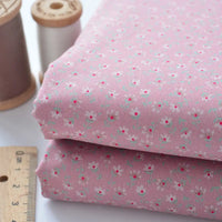 Deux morceaux pliés de Tissu linon de coton fleuri au mètre de Longan Craft France, avec un imprimé floral rose, sont présentés à côté d'une règle en bois et de deux bobines de fil, l'une brune et l'autre blanche.