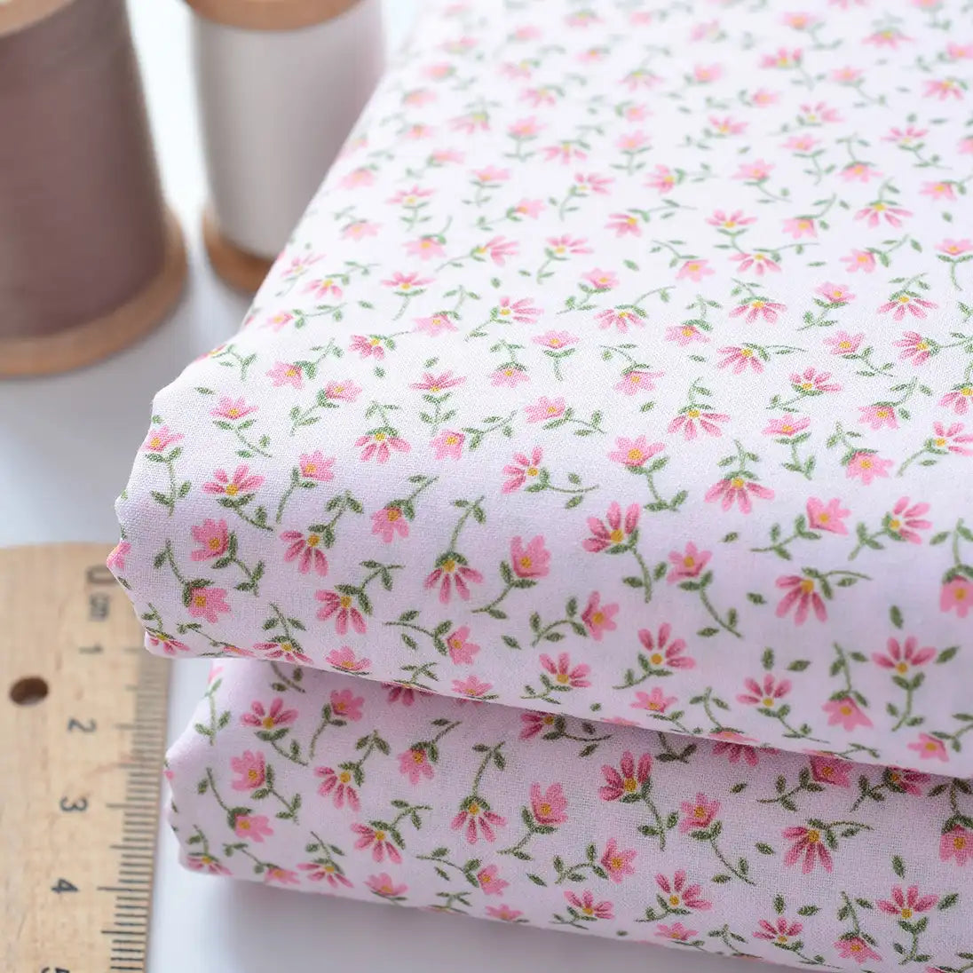 Deux morceaux pliés de Tissu linon de coton fleuri au mètre de Longan Craft France avec un imprimé floral rose sont empilés à côté d'une règle en bois et de deux bobines de fil, marron et blanc, sur une surface blanche.