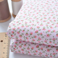 Deux morceaux pliés de Tissu linon de coton fleuri au mètre de Longan Craft France avec un imprimé floral rose sont empilés à côté d'une règle en bois et de deux bobines de fil, marron et blanc, sur une surface blanche.